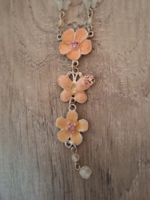 Halsband med blommor och fjäril - Sött halsband med silverfärgad kedja och hängsmycke i form av två blommor och en fjäril i persikotonade nyanser. Blommorna har rosa stenar i mitten och fjärilen är dekorerad med små rosa stenar. Några pärlliknande detaljer längst ner.