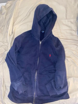 Mörkblå hoodie från Polo Ralph Lauren - Mörkblå hoodie med dragkedja från Polo Ralph Lauren. Klassisk design med huva, fickor framtill och den ikoniska röda loggan broderad på bröstet. Tillverkad i mjukt bomullsmaterial som är skönt att ha på sig.