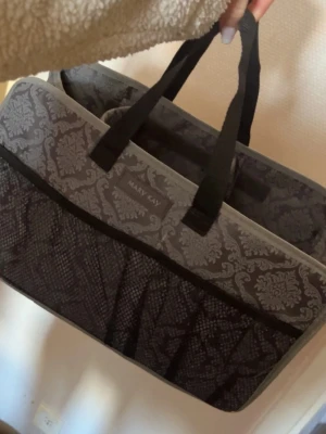 Mary Kay organiseringsväska mönstrad - Snygg och praktisk organiseringsväska från Mary Kay i grått och svart med elegant barockmönster. Väskan har flera fack och meshfickor på utsidan, perfekt för att hålla ordning på smink eller tillbehör. Tillverkad i slitstarkt tyg med två handtag. Går att vika ihop. 