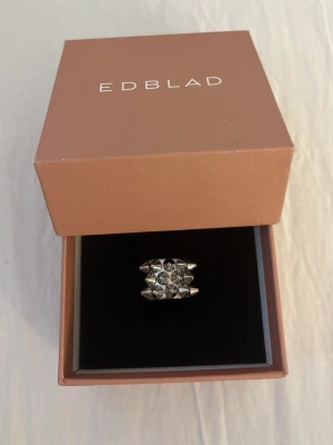 Silvrig ring med nitar från Edblad - Cool och chunky ring i blankt silverfärgat metall från Edblad. Ringen har ett unikt mönster med små nitar och geometriska former på ovansidan som ger en edgy vibe. Perfekt för dig som vill sticka ut med dina accessoarer.