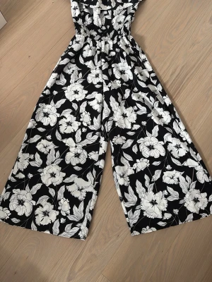 Svartvit blommig jumpsuit från AMISU - Säljer en svartvit jumpsuit med stort blommönster och vida ben. Jumpsuiten har resår i midjan för skön passform och är tillverkad i ett lätt, mjukt tyg som känns luftigt. Perfekt för dig som gillar en chill och trendig stil med mycket mönster. Knappt använd 