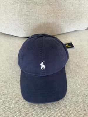 Mörkblå keps från Polo Ralph Lauren - Snygg mörkblå keps från Polo Ralph Lauren med klassisk böjd skärm och vit broderad logga framtill. Kepsen är tillverkad i bomull och har justerbar rem baktill för perfekt passform. Perfekt accessoar för en avslappnad och stilren look.