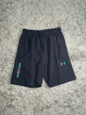 Svarta Under Armour shorts med reflex - Snygga svarta shorts från Under Armour med elastisk midja och dragsko. Loggan och texten 'ARMOUR' i regnbågsfärger på framsidan samt färgglada detaljer på baksidan. Perfekta för träning eller chill. Tillverkade i lätt och bekvämt material. För fler frågor/bilder skriv privat. Hela settet finns i profilen som en egen annons