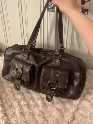 Snygg brun läder väska - Brun läderväska med guldiga detaljer och early 2000’s stil, lite sliten, funkar som handväska eller axelremsväska. Skriv för mer frågor☺️