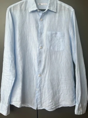 Ljusblå linneskjorta från Zara - Säljer en stilren ljusblå skjorta från Zara i svalt linne. Skjortan har klassisk krage, bröstficka och knappar framtill. Perfekt för varma dagar och enkel att matcha med jeans eller shorts.