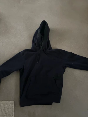 Marinblå hoodie med magficka - En enkel marinblå hoodie ifrån hugo boss med huva och dragsnören. Hoodien har en stor magficka framtill och ribbade muddar vid ärmslut och nederkant. Perfekt för en avslappnad och chill stil. Materialet känns mjukt och bekvämt, aldrig använd pågrund av storleken