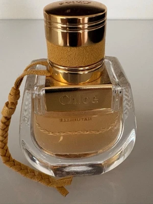 Chloé Nomade Naturelle parfym - Chloé Nomade Naturelle är en elegant parfym i en genomskinlig glasflaska med guldfärgade detaljer och ett flätat band i beige mocka. Flaskan har en exklusiv känsla med rundade former och guldlock. Doften är modern och lyxig, perfekt för dig som gillar stilrena accessoarer.