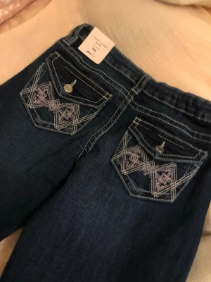 Mörkblå bootcut jeans med brodyr - Snygga mörkblå bootcut jeans med dekorativa fickor bak som har vit och rosa brodyr. Jeansen har kontrastsömmar och vida ben. Jag personligen köper helst dom bootcut jeans som är så flared som möjligt. Perfekta för dig som gillar en retrostil och vill sticka ut lite extra. Materialet är klassisk denim.