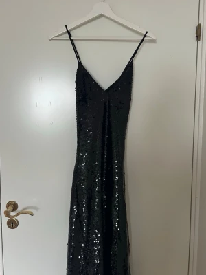 Svart glittrig långklänning - Snygg svart långklänning med smala axelband och v-ringning. Klänningen är täckt av glittriga paljetter som ger en riktigt party-vibe. Klänningen har ett snyggt fall över kroppen och är korsad bak i ryggen. Klänningen är endast använd 1 gång. 