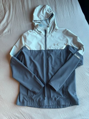 Grå och ljusblå vindjacka Under Armour - Snygg vindjacka från Under Armour i tvåfärgad design med grå nederdel och ljusblå ovandel. Jackan har huva, hel dragkedja framtill och logga på bröstet samt ärmen. Tillverkad i ett lätt och vindtätt material, perfekt för blåsiga dagar.