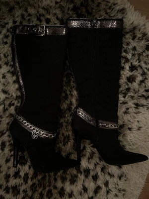 Svarta 2000s höga stövlar  - Säljer mina jätte snygga svarta stövlar med spetsig tå och ormskinnsmönstrade kanter och bälte upptill samt kedjedetalj med ett gulligt hänge vid ankeln ❤️ 