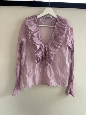 Ljuslila blus med volang från By Ti Mo - Supersöt ljuslila blus från By Ti Mo med djup V-ringning och stora volanger längs halsen. Blusen har långa, lätt plisserade ärmar och är gjord i ett tunt, skirt material som känns luftigt och trendigt.