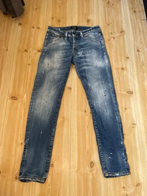 Jack and Jones jeans - Säljer ett par blå jeans med slitna och tvättade detaljer samt små färgstänk för en trendig grisch stil. Slim fit storlek 30/32 