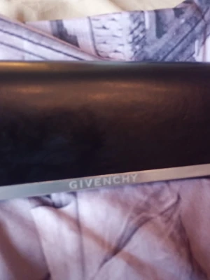 Svart solglasögon fodral från Givenchy - Snyggt solglasögon fodral från Givenchy med stilren silverfärgad kant och Givenchy-logga framtill. Den är av slätt läder och har en klassisk, avlång form. Perfekt för dig som vill ha en elegant accessoar med lyxig känsla.