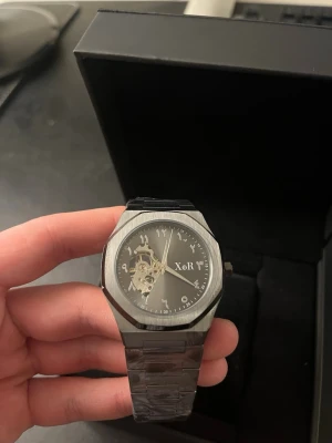 XoR Obsucra 41 mm Automatiskt urverk HELT NY - Snygg svart klocka från XoR watches med stainless steel armband och grå/svart urtavla. Klockan har ett öppet urverk som syns genom urtavlan och arabiska siffror i en unik design. Skickas med XoR original box.