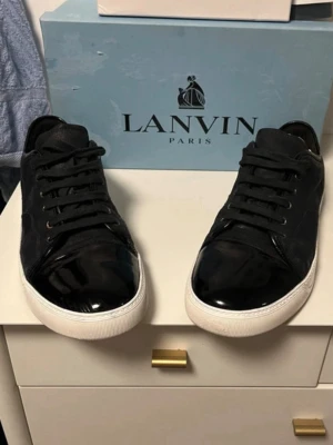 Svarta Lanvin sneakers med lackdetalj - Snygga svarta sneakers från Lanvin med klassisk låg profil. Skorna har en unik blank lackdetalj på tån och vit sula som kontrast. Tillverkade i en mix av mocka och läder, perfekta för dig som vill ha en stilren men ändå lyxig sneaker.