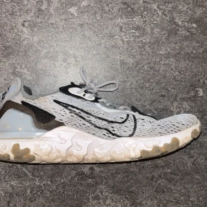 Nike React Vision vita sneakers - Nike React Vision sneakers i vitt och grått med svart swoosh och detaljer. Ovandelen är i mesh för bra ventilation och sulan är tjock och mönstrad för extra komfort. Skorna har en modern, sportig look med snörning och reflekterande detaljer.