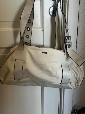 Beige handväska från fiorelli - Cool beige handväska från fiorelli med bred axelrem och metallringar som detaljer. Väskan har en mjuk och rymlig form, med svarta kanter och dekorativa remmar på framsidan. Tecken på användning 