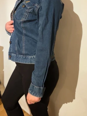 Klassisk blå jeansjacka - Snygg blå jeansjacka med klassisk passform och detaljer som bröstfickor med lock och knappar framtill. Jackan har justerbar midja baktill och markerade sömmar för en tidlös look. Perfekt att slänga över en hoodie eller t-shirt.