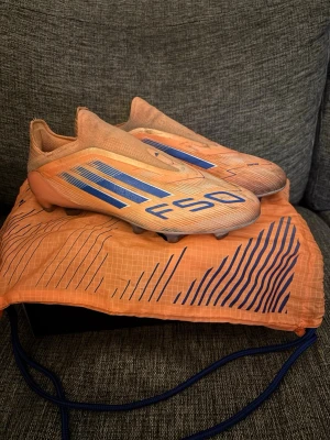 Adidas F50 orange/blå fotbollsskor - Säljer ett par Adidas F50 fotbollsskor i orange med blå detaljer och logga. Skorna har en strömlinjeformad design utan snörning och en lätt ovandel i syntetmaterial. Sulan har fasta dobbar för spel på naturgräs. Medföljer matchande orange påse med blått mönster. Skorna är andvända i ungefär en månad och är i väldigt bra skick utan några som helst skador eller defekter, dom är lite smutsiga min ingeting mer än det allt og medföljer!