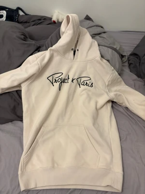 Beige hoodie från Project X Paris - Snygg beige hoodie från Project X Paris med svart broderad logga på bröstet. Hoodien har stor känguruficka framtill, ribbade muddar och en klassisk huva med dragsko. Perfekt för en avslappnad streetwear-look.