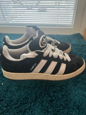 Adidas Campus 00 svarta  - Klassiska Adidas Campus sneakers i svart mocka med vita detaljer och de ikoniska tre ränderna på sidan. Skorna har vita skosnören och Adidas logga på plösen. Låg modell med vit sula som ger en clean och tidlös look.