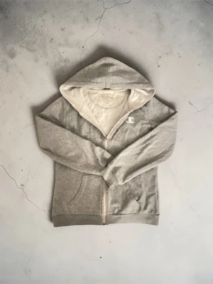 Grå zip-hoodie från Champion - Grå hoodie med dragkedja från Champion. Klassisk modell med huva, fickor framtill och ribbade muddar. Mjuk insida och diskret logga på bröstet. Perfekt för en chill och sportig stil. Det är bara att höra av er vid frågor! Med vänlig hälsning, Valter 😊