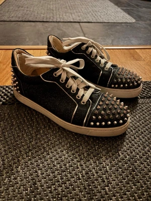 Christian Louboutin sneakers med nitar - Svarta sneakers från Christian Louboutin med vita snören och vita detaljer. Skorna har silvriga nitar över tå och häl, samt en vit sula. Materialet är glittrigt tyg som ger en cool och edgy vibe. Perfekta för dig som vill sticka ut med din stil.