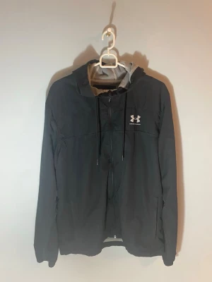 Svart vindjacka från Under Armour - Snygg svart vindjacka från Under Armour med vit logga på bröstet. Jackan har huva med meshfoder, dragkedja framtill och justerbara snören vid huvan. Perfekt för blåsiga dagar och sportiga outfits.