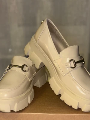 Beige chunky loafers med metallspänne - Stilrena beige loafers med chunky sula och dekorativt metallspänne framtill. Skorna har en modern, robust design och är tillverkade i glansigt material som ger en fräsch look. Har endast använts 2 gånger, därav den smutsiga sulan :) Lite smuts och repor förekommer, har försökt att få med dessa på bild. Inget som märks av så ☺️ Köpt på Zalando för några år sen, skolådan följer med!! 