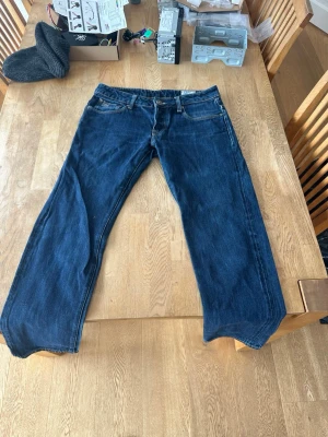 Mörkblå raka jeans från g-star - Säljer ett par klassiska mörkblå jeans från Lee med rak passform och fem fickor. Jeansen har kontrastsömmar och en läderpatch med logga bak i midjan. Tillverkade i slitstark denim, perfekta för en avslappnad och tidlös stil.