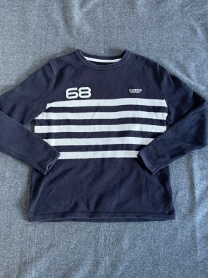Mörkblå stickad tröja Morris - Snygg mörkblå stickad tröja från Morris med vita horisontella ränder och siffran 68 på bröstet. Tröjan har rund halsringning och långa ärmar. Perfekt för dig som gillar klassisk men sportig stil.