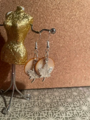 Handgjorda örhängen med färgade snäckskals pärlor och löv - Handgjorda örhängen med orange färgade snäckskal och berlock. Hängande modell med krokar i metall. Perfekta för dig som gillar naturinspirerade accessoarer och vill sticka ut. Inte nickelfria 
