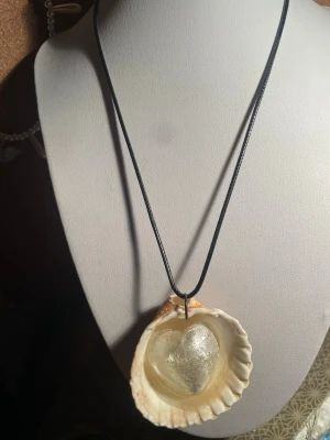 Handgjort halsband med snäcka  - Unikt handgjort halsband med svart snöre och stort hänge av en naturlig snäcka. Inuti snäckan finns ett genomskinligt hjärta med skimrande silverfärg. Perfekt för dig som gillar strandinspirerade accessoarer och vill sticka ut med något annorlunda.