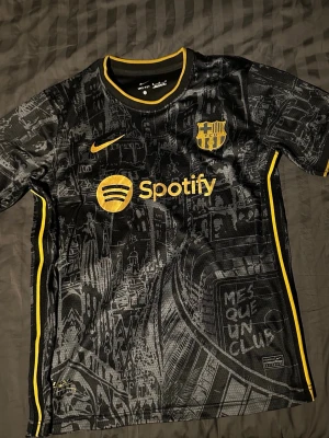 FC Barcelona svart matchtröja Nike fotbollströja - Ej använd. Snygg svart FC Barcelona matchtröja från Nike med gula detaljer och tryck. Tröjan har klubbmärke, Nike-logga och Spotify som sponsor framtill. Unikt mönster med Barcelonas stadssilhuett och texten 'Mes Que Un Club'. Perfekt för fotbollsfans!