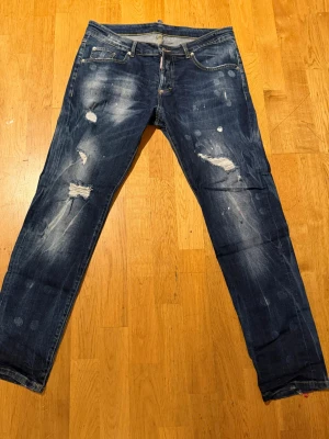 Dsquared2 jeans  - Säljer ett par blå jeans från Dsquared2 med slitna detaljer och trasiga partier. Jeansen har klassisk femficksdesign, gul kontrastsöm och en smal passform. Materialet är denim och det finns en röd Dsquared2-logga vid gylfen.