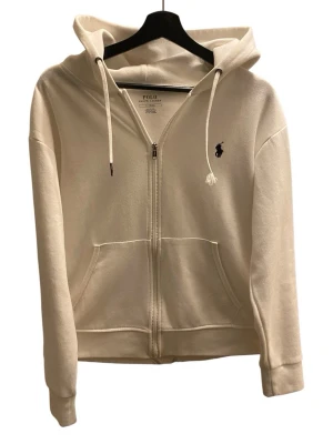 Vit hoodie från Polo Ralph Lauren - Beige hoodie med dragkedja från Polo Ralph Lauren. Klassisk design med huva, dragsnören och kängurufickor framtill. Liten broderad logga i svart på bröstet. Tillverkad i mjukt bomullsmaterial för en skön och avslappnad look.