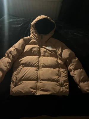 Beige pufferjacka från The North Face - Säljer en beige pufferjacka från The North Face med huva och dragkedja framtill. Jackan har två sidofickor med dragkedja och logga på bröstet samt ryggen. Perfekt för kalla vinterdagar och riktigt skön att bära. Man kan vända den så den blir svart men det får man välja själv. Nypris 3000