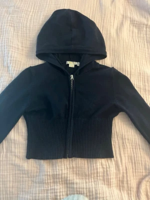 Mörkblå cro-ribbad hoodie med dragkedja - Säljer en mörkblå hoodie med dragkedja framtill och ribbad midja. Hoodien har en croppad passform och långa ärmar. Materialetone är mjukt och stretchigt, perfekt för en chill och stilren look. Klassisk huva och minimalistisk design utan synliga loggor.