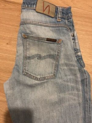 Ljusblå jeans från Nudie Jeans - Säljer ett par ljusblå jeans från Nudie Jeans med klassisk femficksmodell och sny Parlour-våg på bakfickan. Jeansen har slitningar clock washed look och är tillverkade i bomull. Perfekta för dig som gillar avslappnad stil. W30 L32