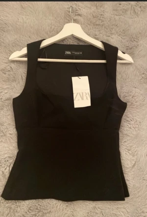 Svart ärmlös topp från Zara - Snygg svart ärmlös topp från Zara med breda axelband och figurnära passform. Toppen har en stilren design och lätt utsvängd nederdel, perfekt att matcha med jeans eller kjol. Materialet känns mjukt och stretchigt.