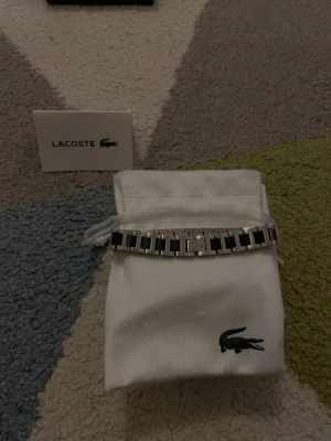 Lacoste silverfärgad armbandsklocka - Snygg armbandsklocka från Lacoste med länkarmband i silverfärgad metall och svarta detaljer. Klockan kommer med originalpåse och box med den klassiska krokodilloggan. Perfekt accessoar för dig som gillar stilrena och moderna detaljer.