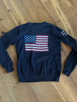 Mörkblå stickad tröja från GANT - Snygg mörkblå stickad tröja från GANT med amerikansk flagga på bröstet och broderad text på ärmen. Tröjan har rund halsringning och ribbade muddar vid ärmslut och nederkant. Perfekt för dig som gillar klassisk och sportig stil.