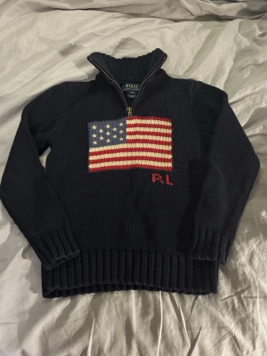 Mörkblå stickad tröja Polo Ralph Lauren S(8) - Mörkblå stickad tröja från Polo Ralph Lauren med halv dragkedja och ribbade muddar. På bröstet finns en stor amerikansk flagga i rött, vitt och blått samt RL-broderi. 