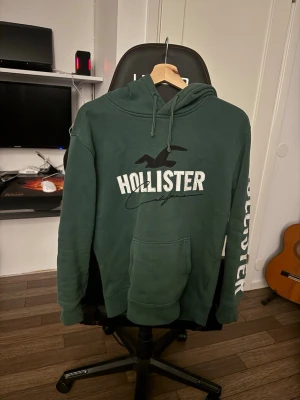 Grön Hollister hoodie med tryck - Mörkgrön hoodie från Hollister med stor logga och text på bröstet samt längs ena ärmen. Klassisk känguruficka framtill och justerbar huva med snören. Perfekt för en avslappnad och cool stil. Mjuk bomullsblandning för extra komfort. Den är xs men passar mig som är s super bra 
