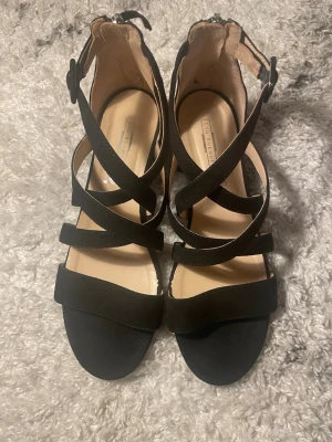 Svarta sandaler med klack - Säljer ett par snygga svarta sandaler med korsade remmar och justerbar spänne runt ankeln. Skorna har en låg blockklack och är tillverkade i mockaliknande material med ljus innersula. Perfekta för dig som gillar stilrena och trendiga skor. Bara använt en gång!