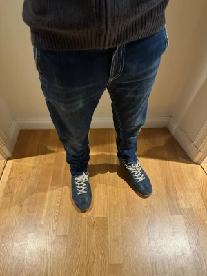 Mörkblå jeans från dondup - Säljer ett par mörkblå jeans från dondup med vita kontrastsömmar och coola detaljer som dragkedjor på bakfickorna. Jeansen har en avslappnad passform och är perfekta för en streetwear-look.