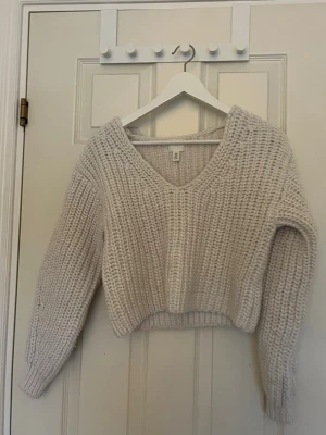 Beige stickad tröja från H&M - Mysig beige stickad tröja från H&M med grovstickat mönster och djup v-ringning. Tröjan har långa ärmar och en något kortare, boxig passform. Perfekt att slänga på sig när det är kyligt ute.