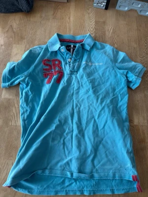 Blå pikétröja från SR Racing - Snygg blå pikétröja från SR Racing med röd broderad SR77-detalj på bröstet och vit logga. Klassisk krage och korta ärmar, perfekt till sommaren. Röd kontrastsöm i kragen och liten detalj vid nederkanten. Skön och luftig bomullskvalitet.