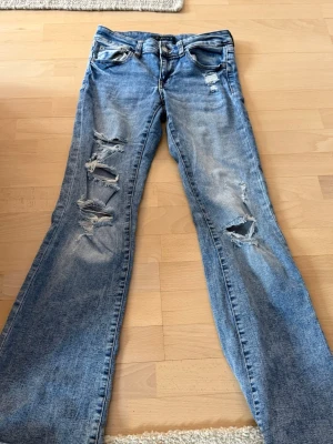 Blå slitna jeans från Aeropostale - Säljer ett par blå jeans från Aeropostale med flera slitningar och hål på benen för en riktigt trendig och avslappnad look. Jeansen har klassisk femficksmodell och lätt utsvängda ben. Perfekta för dig som gillar streetstyle och en edgy vibe.
